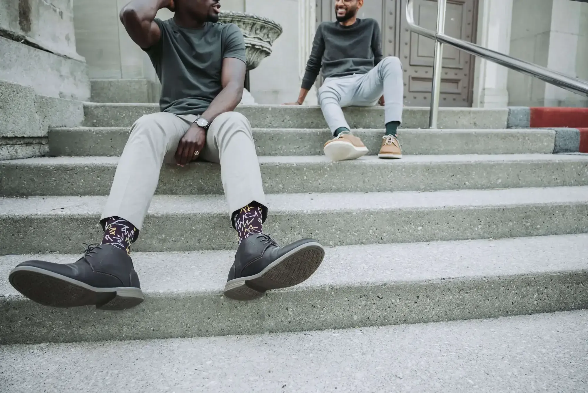 Two men sitting on stone steps, one wearing patterned dress socks with dark boots